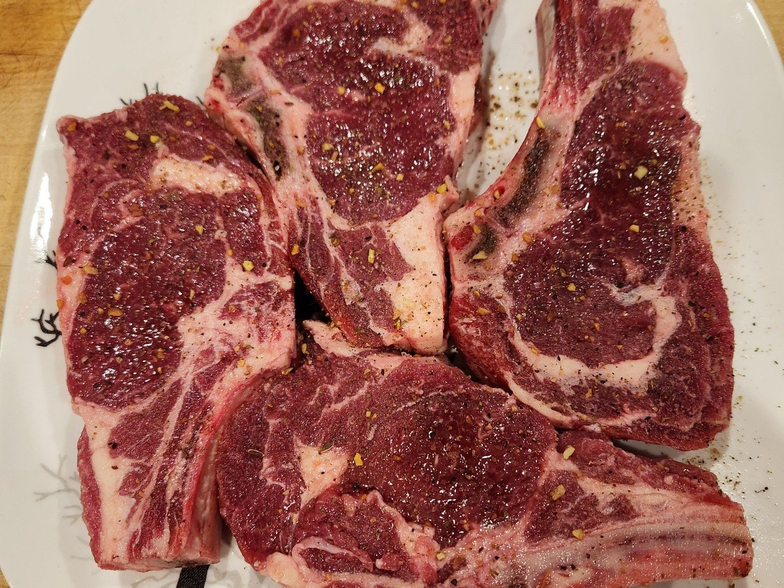 Four seasoned ribeye steaks arranged on a white plate, showcasing rich marbling and a sprinkle of black pepper and spices, ideal for grilling or pan-searing.