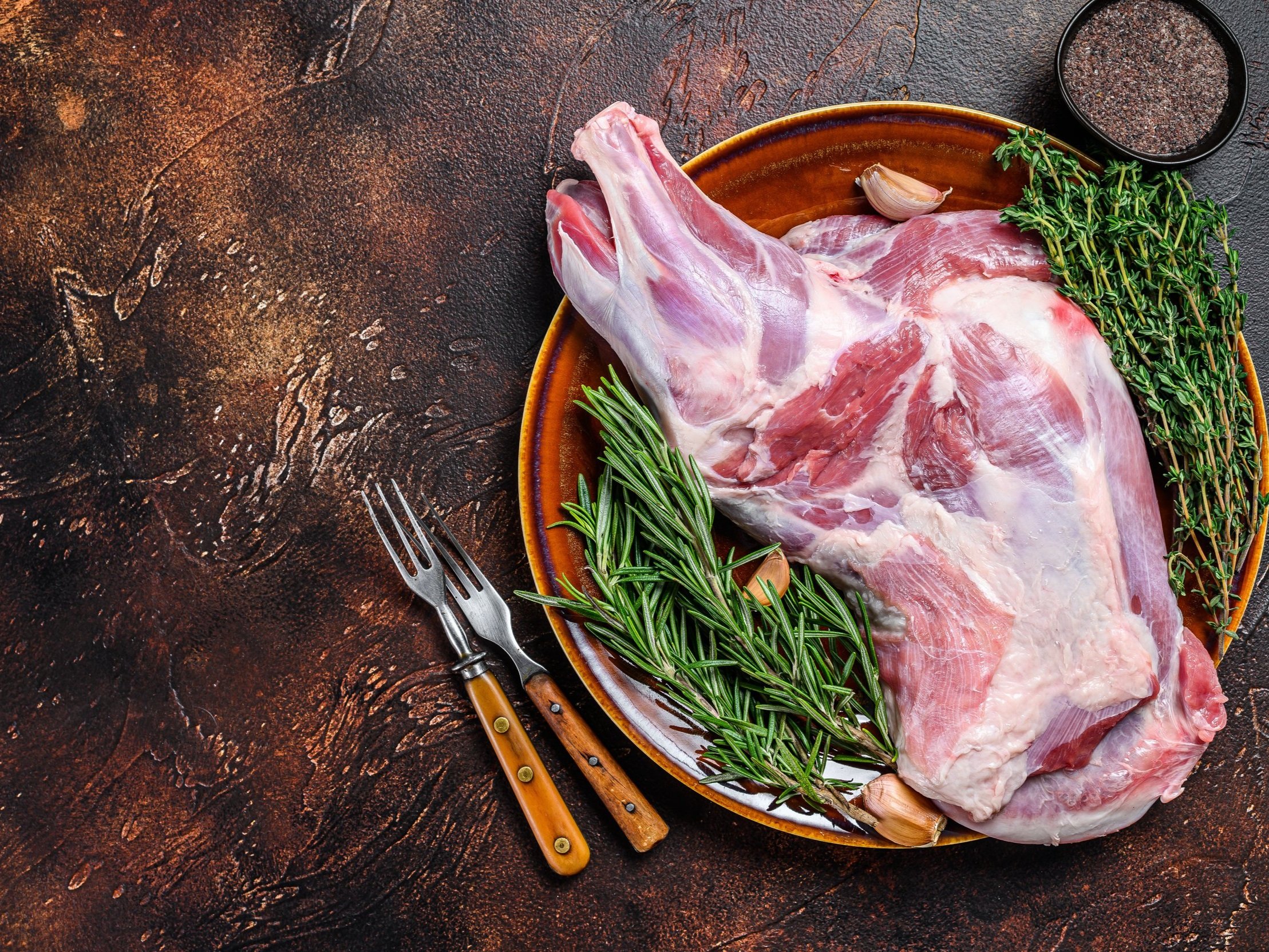 Raw lamb leg on a brown plate surrounded by fresh rosemary, thyme, and garlic cloves, with two forks and a bowl of black pepper on a textured dark surface.