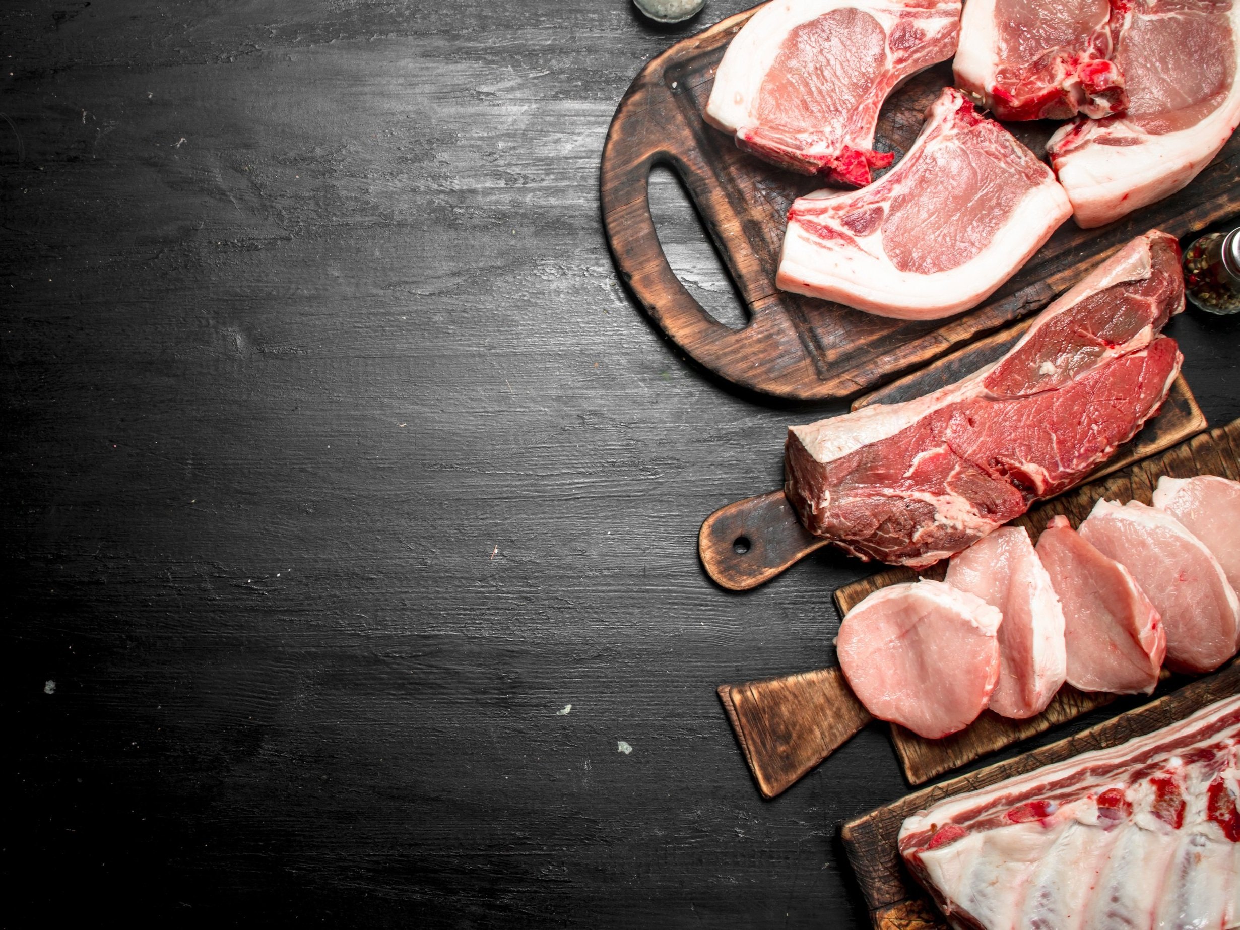 Assorted cuts of raw meat displayed on wooden cutting boards, including pork chops, beef steaks, and ribs, set against a dark textured background. Perfect for culinary and food preparation themes.
